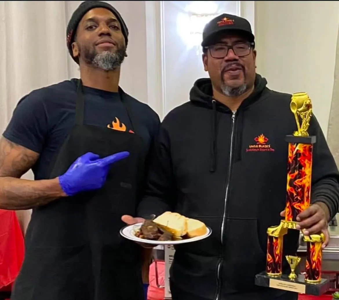 Two cook-off winners with a trophy and a plate of Pepperpot with bread